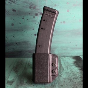 May include: A black magazine and magazine holder. The magazine is inserted into the holder. The holder is attached with two screws. The background is a teal color with paint splatters.