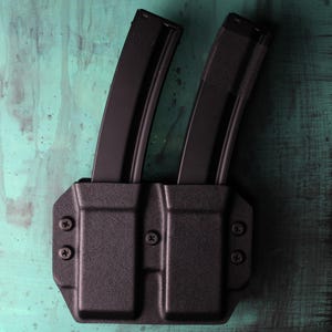May include: Black double magazine carrier with two black magazines inserted. The carrier is made of a textured material and is secured with screws. The magazines are upright and appear to be for a firearm. The background is a distressed teal color.