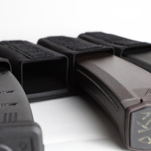 May include: Close-up of several black magazine pouches with a dark brown magazine. The pouches have a rectangular shape with a velcro closure. The magazine is dark brown with a textured surface.
