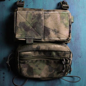 May include: Camouflage pattern chest rig with two pouches. The top pouch has a square velcro patch and the bottom pouch has a zipper closure. The rig is made of a durable fabric and features adjustable straps and buckles. The color scheme is a mix of green, brown, and gray.