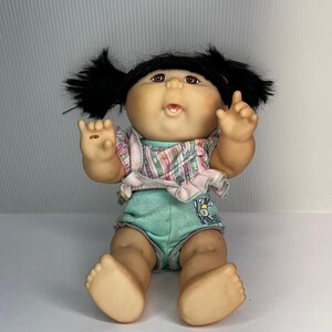 May include: A vintage doll with a round face, black pigtails, and brown eyes. The doll wears a patterned top with pink trim and light blue shorts with a bunny graphic. The doll is in a sitting position with its arms raised.