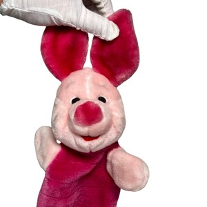 May include: A plush Piglet hand puppet in shades of pink and red. The puppet has large, floppy ears, a round snout, and a smiling expression. The puppet is held by a gloved hand against a white background.