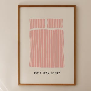 Puede incluir: Un estampado artístico enmarcado con una ilustración minimalista de una cama con ropa de cama rosa y el texto "LET'S STAY IN BED". La obra de arte se muestra sobre una cama con almohadas y ropa de cama a rayas, creando una escena de dormitorio acogedora.