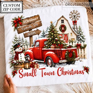 May include: A white t-shirt featuring a graphic of a red pickup truck, a snowman, a barn, and a Christmas tree. The design includes a wooden sign reading Christmas, Fayetteville, TN, 37334 and the red text Small Town Christmas at the bottom.