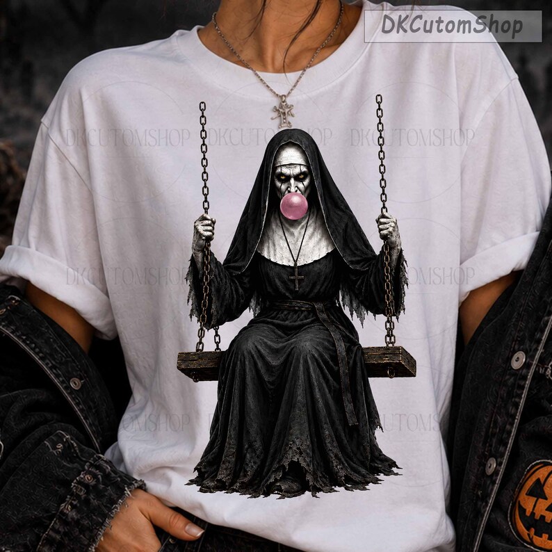 May include: A white t-shirt featuring a graphic of a nun in a black habit sitting on a swing suspended by chains. The nun has a pale face and is blowing a large pink bubble with bubblegum. A silver cross necklace hangs around the nun's neck.