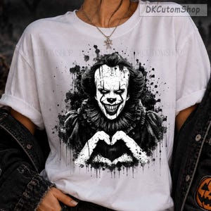 May include: A white t-shirt featuring a black and white graphic print of a clown with sharp teeth and a ruffled collar, making a heart shape with hands. The design includes ink splatter effects and the text DKCutomShop in the top right corner.