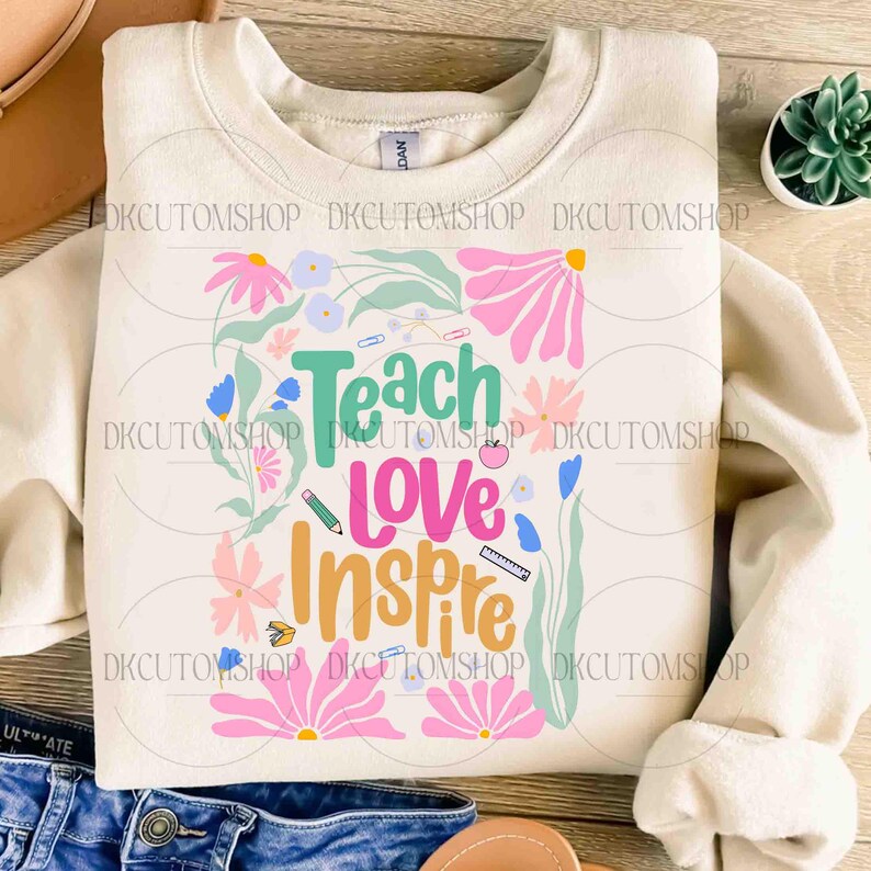 May include: A beige sweatshirt with a floral design and the text "Teach Love Inspire" in colorful letters. The design includes a pencil, a ruler, an apple, and other school-related items.