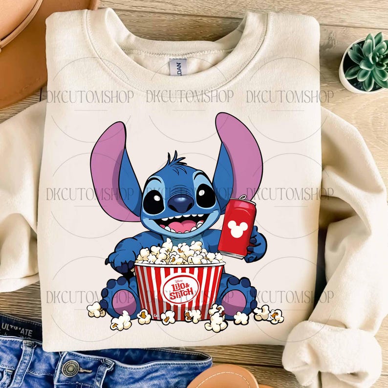 Puede incluir: Una sudadera de color crema con una imagen de dibujos animados de Stitch de Lilo & Stitch. Stitch es azul con orejas grandes, sosteniendo una lata roja con un logotipo de Mickey Mouse y una cubeta de palomitas de ma&iacute;z. La cubeta de palomitas tiene el texto "Lilo & Stitch".