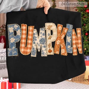 May include: A black t-shirt featuring the word PUMPKIN in large, fabric-appliqué letters. Each letter displays a different pattern, including blue floral, orange gingham, and tan with a small pumpkin and vine graphic on the letter I.