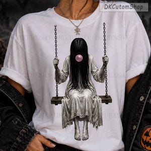 May include: A white short-sleeved t-shirt featuring a graphic print of a figure with long black hair wearing a light-colored dress, sitting on a swing suspended by chains, blowing a pink bubble. The text DKCutomShop appears in the corner.
