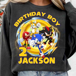 May include: A black sweatshirt with a graphic of Sonic the Hedgehog and his friends. The text "Birthday Boy" is above the graphic and the number "2" and the name "Jackson" are below the graphic.