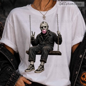 May include: A white t-shirt featuring a graphic print of a hockey-masked figure in a dark jumpsuit sitting on a swing, blowing a pink bubble. The figure wears dark sneakers and makes a peace sign with one hand. The design is centered on the chest.