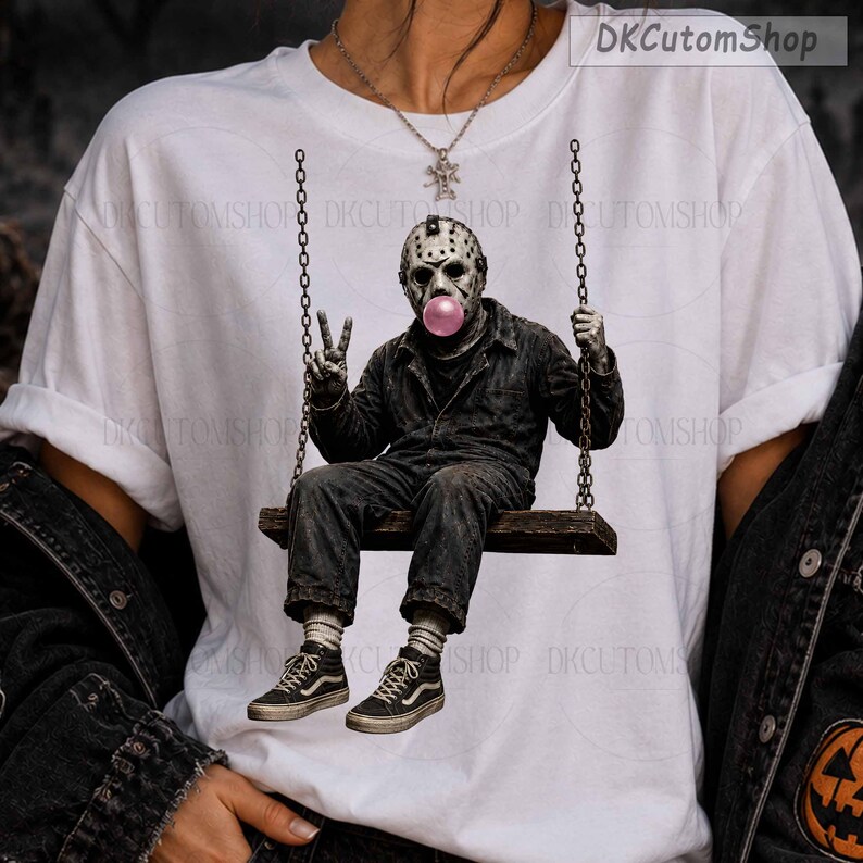 May include: A white t-shirt featuring a graphic print of a hockey-masked figure in a dark jumpsuit sitting on a swing, blowing a pink bubble. The figure wears dark sneakers and makes a peace sign with one hand. The design is centered on the chest.
