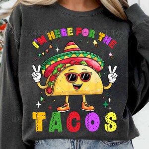 May include: A dark gray sweatshirt with the text "I'm here for the tacos" in colorful letters. A cartoon taco wearing a sombrero and sunglasses is holding up two fingers in a peace sign.