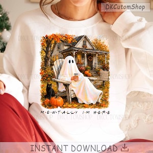 Op de afbeelding: Een wit sweatshirt met ronde hals en een grafische print van een geest die op een houten stoel op een veranda zit en een koffiebeker vasthoudt. Een zwarte kat en oranje pompoenen zijn zichtbaar. De tekst MENTALLY I'M HERE staat onder de afbeelding.