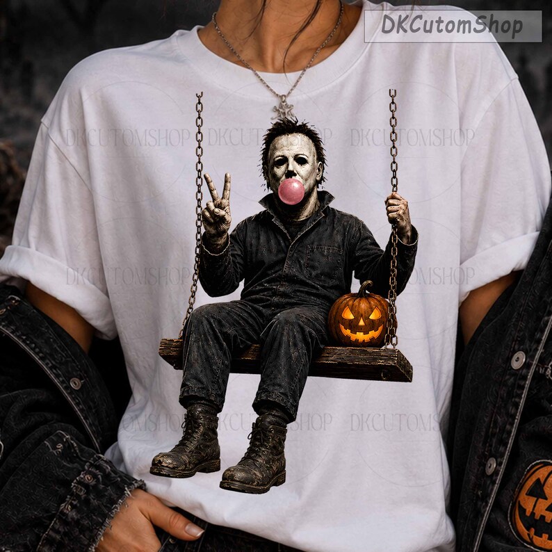 May include: A white t-shirt featuring a graphic of a character in a dark jumpsuit sitting on a swing, blowing a pink bubble, and making a peace sign. A glowing orange jack-o'-lantern sits on the swing next to the character. The text DKCutomShop appears.