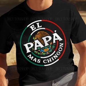 Puede incluir: Camiseta negra con un dise&ntilde;o circular. El texto "EL PAP&Aacute; MAS CHINGON" est&aacute; en blanco, verde y rojo. El dise&ntilde;o incluye un emblema de &aacute;guila marr&oacute;n y un borde verde y blanco, que representa la bandera mexicana.
