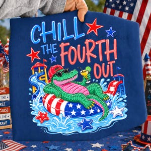 May include: Navy blue graphic tee with the text "CHILL THE FOURTH OUT". The design features a cartoon alligator wearing sunglasses and a red hat, relaxing on a patriotic inner tube with a cocktail. Red and blue stars and water slides complete the festive look.