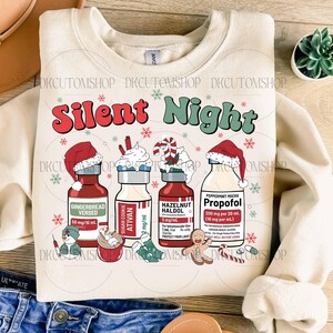 Op de afbeelding: Cr&egrave;mekleurige sweatshirt met de woorden "Silent Night" in rood en groen. Het ontwerp bevat illustraties van flessen met feestelijke namen zoals "Gingerbread Versed" en "Peppermint Mocha Propofol", samen met sneeuwvlokken en peperkoekmannetjes.