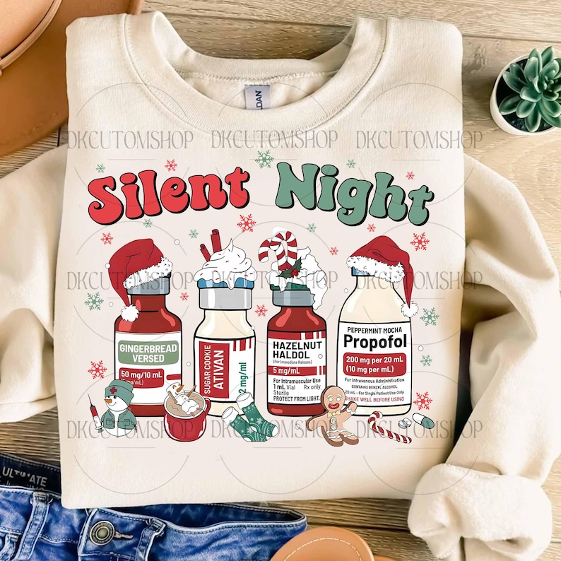 Op de afbeelding: Cr&egrave;mekleurige sweatshirt met de woorden "Silent Night" in rood en groen. Het ontwerp bevat illustraties van flessen met feestelijke namen zoals "Gingerbread Versed" en "Peppermint Mocha Propofol", samen met sneeuwvlokken en peperkoekmannetjes.
