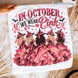 以下が含まれることがあります： IN OCTOBER WE WEAR PINKというテキストがプリントされた白いTシャツ。ピンクの魔女の帽子とフリルのついたピンクのドレスを身にまとった5人の人物が描かれており、ピンクのリボン、小さなハート、星、黒いコウモリのモチーフが添えられています。