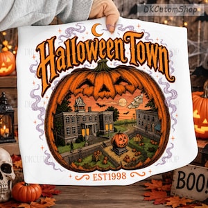 May include: A white fabric item featuring a large orange jack-o'-lantern graphic with a detailed town scene inside. The text reads Halloween Town and EST 1998 in orange lettering. Purple smoke swirls around the pumpkin, with stars and a crescent moon.