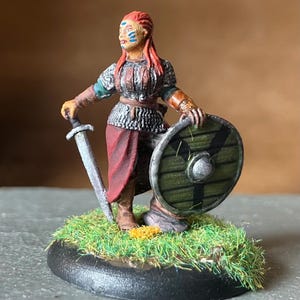 Könnte beinhalten: Miniaturfigur einer Kriegerin, die ein Schwert und einen Schild hält. Sie trägt Kettenrüstung, einen roten Rock und hat rotes Haar. Der Schild ist grün mit schwarzen Streifen. Die Figur steht auf einem Sockel mit Kunstgras.