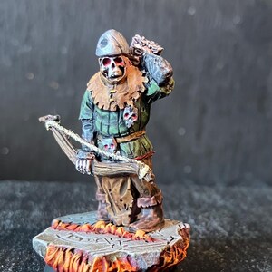 Può includere: Figurina in miniatura di un arciere scheletrico, dipinta con dettagli. La figura indossa una tunica verde, pantaloni marroni e un elmo. Tiene un arco e una freccia, con una faretra sulla schiena. La base è una piattaforma di pietra con un design infuocato.