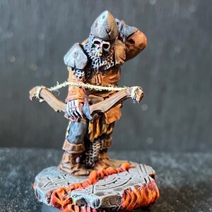 Könnte beinhalten: Miniaturfigur eines Skelettkriegers in brauner und grauer Rüstung, der einen Bogen hält. Die Figur steht auf einem Steinsockel mit feurigen orangefarbenen Akzenten. Der Schädel des Kriegers ist sichtbar, und die Details sind fein bemalt.