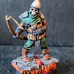 Könnte beinhalten: Miniaturfigur eines Skelettbogenschützen mit detaillierter Rüstung und Helm. Die Figur steht auf einem Steinsockel mit einem feurigen, lavaartigen Effekt. Der Bogenschütze hält einen Bogen und Pfeil, bereit zum Schießen.