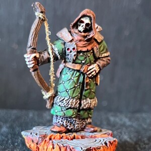 Könnte beinhalten: Miniaturfigur eines Skelettbogenschützen in einer Kapuzenrobe, der einen Bogen hält. Die Figur ist in Grün-, Braun- und Beigetönen bemalt und steht auf einem Sockel mit einem feurigen Design. Detaillierte Fantasy-Figur.