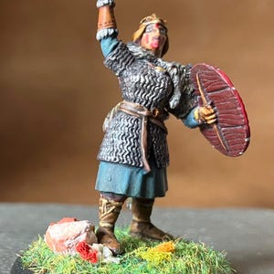 Könnte beinhalten: Miniaturfigur eines Kriegers in Kettenrüstung, der einen Hammer hochhält und einen Schild. Die Figur trägt einen blauen Tunic und braune Stiefel. Der Sockel zeigt grünes Gras und einen kleinen Felsen mit roten und weißen Details. Historisches oder Fantasy-Thema.