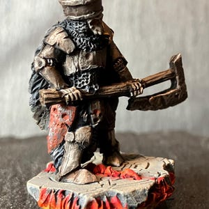 Könnte beinhalten: Miniaturfigur eines mittelalterlichen Kriegers in Rüstung, der eine große Axt hält. Die Figur ist in metallischen Tönen mit einem rot-weißen Schilddetail bemalt. Der Sockel zeigt eine Steinplattform mit Flammen.