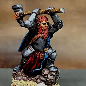 Könnte beinhalten: Miniaturfigur eines Zwergenkriegers in metallgrauer Rüstung, der einen Hammer schwingt. Der Zwerg hat einen roten Bart und ist in Kampfhaltung dargestellt. Die Rüstung hat rote und blaue Akzente. Der Sockel ist strukturiert mit einem Schädel.