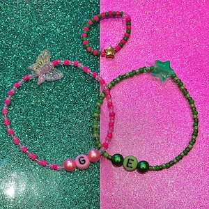 May include: Three beaded bracelets with pink and green beads. One bracelet has a butterfly charm, another has a star charm, and the third has letter charms that spell "G E".