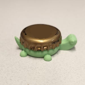 May include: A light green turtle-shaped bottle opener with a gold-colored bottle cap as its shell. The turtle has small legs and a head, all in a light green color. The bottle cap has text and a textured edge.