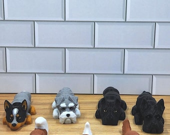 May include: A collection of eight small, 3D printed dog figurines. The figurines are in various breeds, including a husky, a schnauzer, a cavalier king charles spaniel, and a labrador retriever. The figurines are all in a lying down position and are arranged in a row on a wooden surface.
