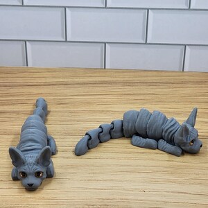 May include: Two gray, articulated, sphinx cat figurines. The cats are posed in different positions, one is lying down and the other is sitting up.