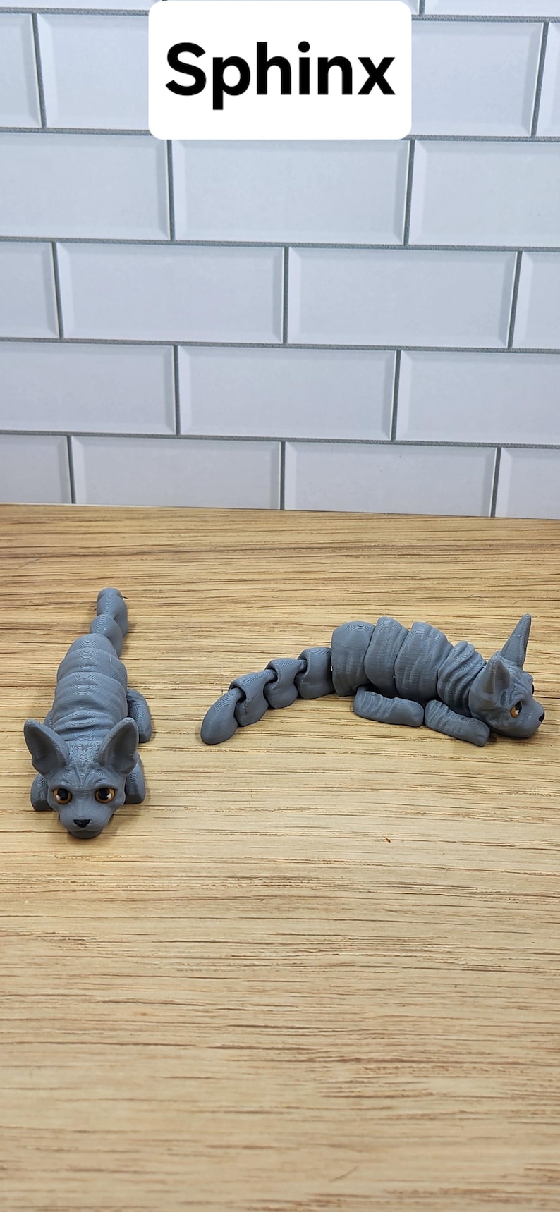May include: Two gray, articulated, sphinx cat figurines. The cats are posed in different positions, one is lying down and the other is sitting up.