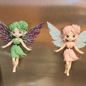 May include: Two fairy figurines with pointed ears and floral hair accessories. The left fairy has a green dress and dark purple glitter wings. The right fairy has a light pink dress and pale blue wings. Both figures have large eyes and are posed as if flying.