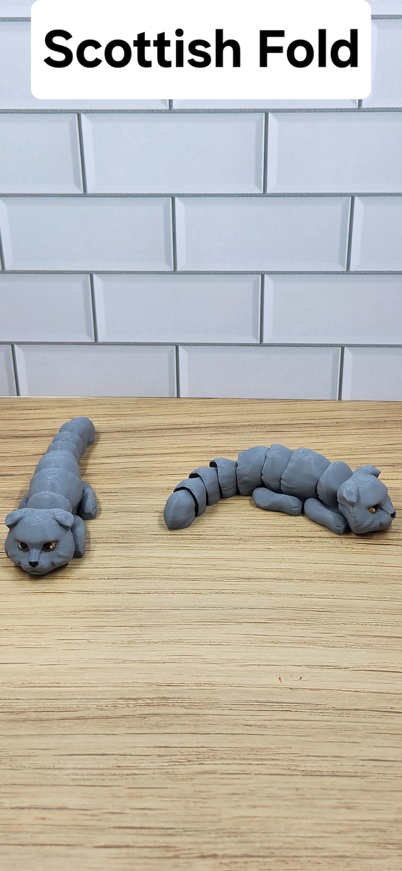 May include: Two gray, segmented, articulated, 3D printed Scottish Fold cat figurines. The cats are posed in a lying down position.