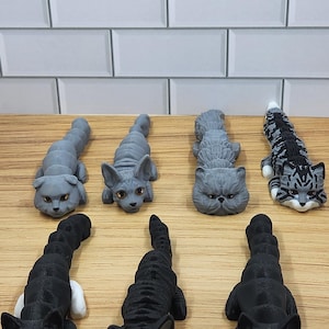May include: Seven 3D printed cat figurines in various poses. The cats are all gray or black with white markings. The figurines are arranged on a wooden surface.