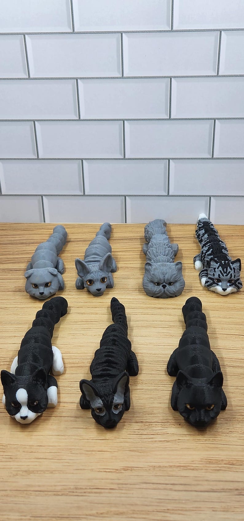 May include: Seven 3D printed cat figurines in various poses. The cats are all gray or black with white markings. The figurines are arranged on a wooden surface.