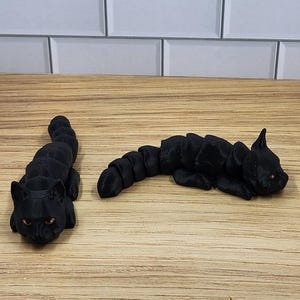 May include: Two black 3D printed cat figurines, one lying down and one sitting up, on a wooden surface.