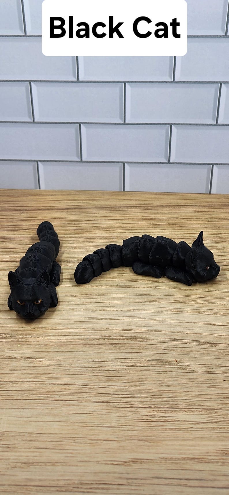 May include: Two black 3D printed cat figurines, one lying down and one sitting up, on a wooden surface.