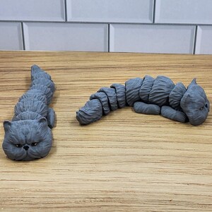 May include: Two gray, 3D printed, Persian cat figurines. The cats are lying down with their heads resting on the surface. The cats are on a wooden surface.