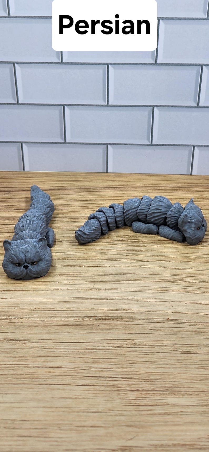May include: Two gray, 3D printed, Persian cat figurines. The cats are lying down with their heads resting on the surface. The cats are on a wooden surface.