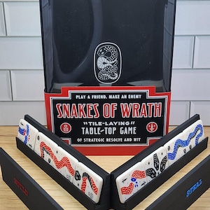 May include: A Snakes of Wrath tile-laying table-top game box with a black and red design. The box features a white snake logo and the text Play a Friend, Make an Enemy. Two black tile racks hold white tiles with red, black, and blue snake patterns.