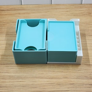 May include: A teal plastic card holder with two compartments and a white plastic base. The left compartment is open, while the right compartment contains a stack of teal cards. The white base features a series of small vertical slots along the side.