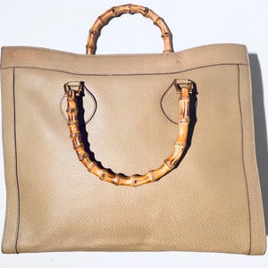 May include: A beige leather tote bag with bamboo handles. The bag has a rectangular shape and a top zipper closure.